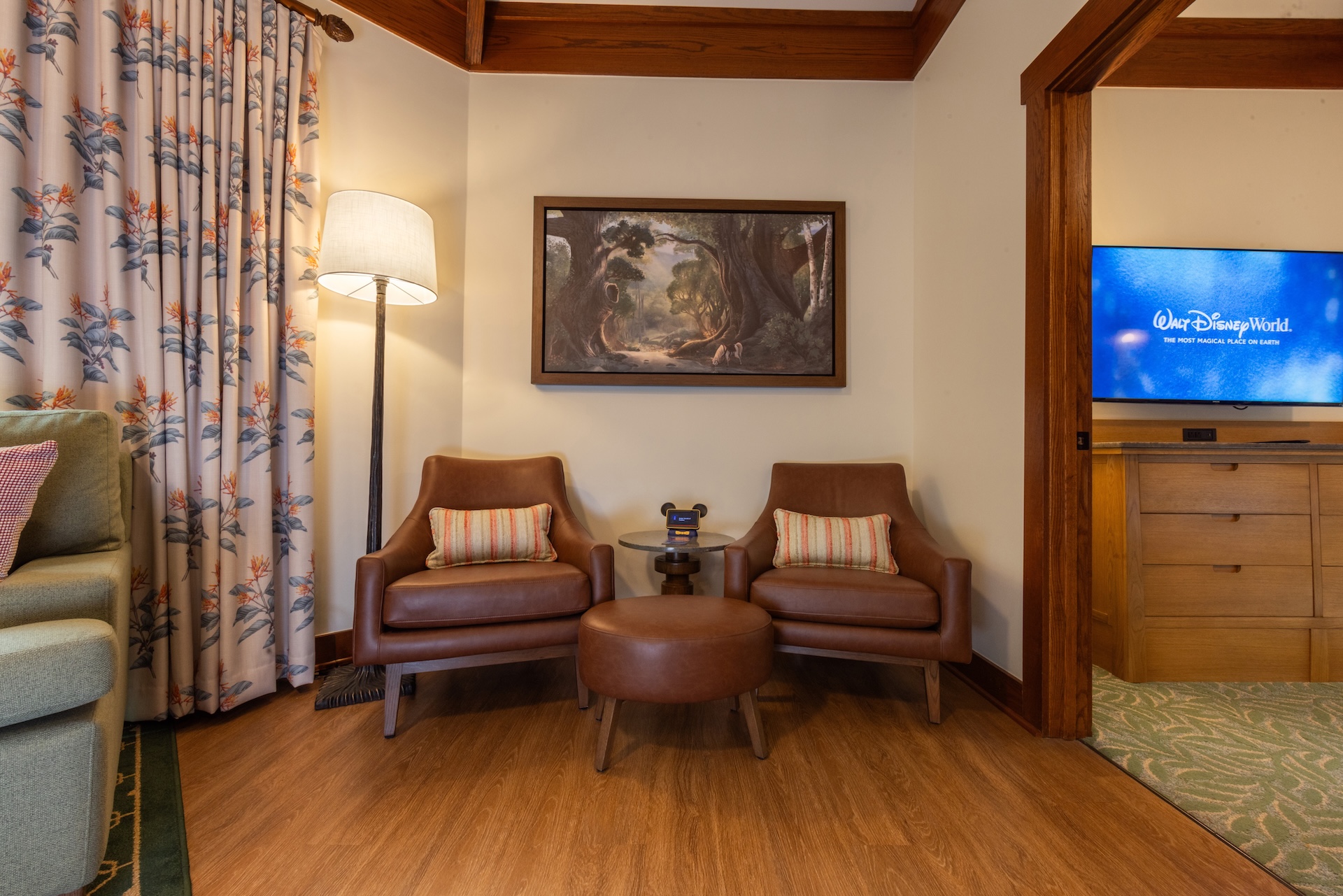 Two Brown Leather Chairs with a Round Ottoman Between Them in a Cozy Lounge with a Side Table and Lamp and a Forest Painting on the Wall