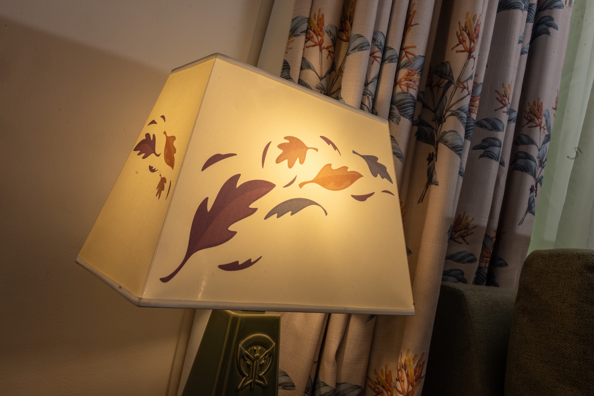 Angled Beige Lampshade with a Leaf Pattern Casting Warm Light Beside Botanical Curtains in a Living Room
