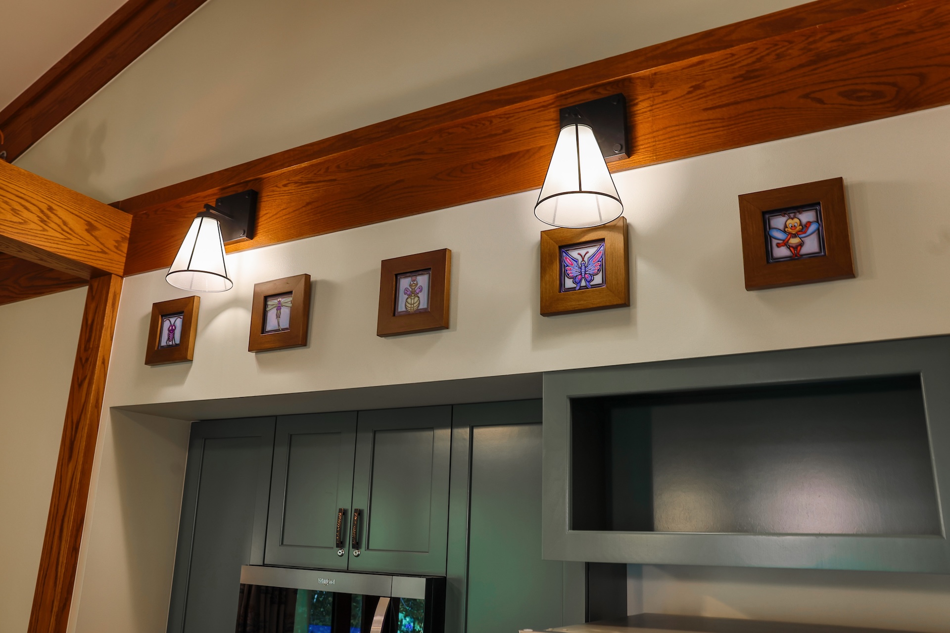 Kitchen Area with Gray green Cabinets Wooden Beam and Two Cone shaped Wall Lights Small Wood framed Insect Pictures Line the Wall Above the Cabinets