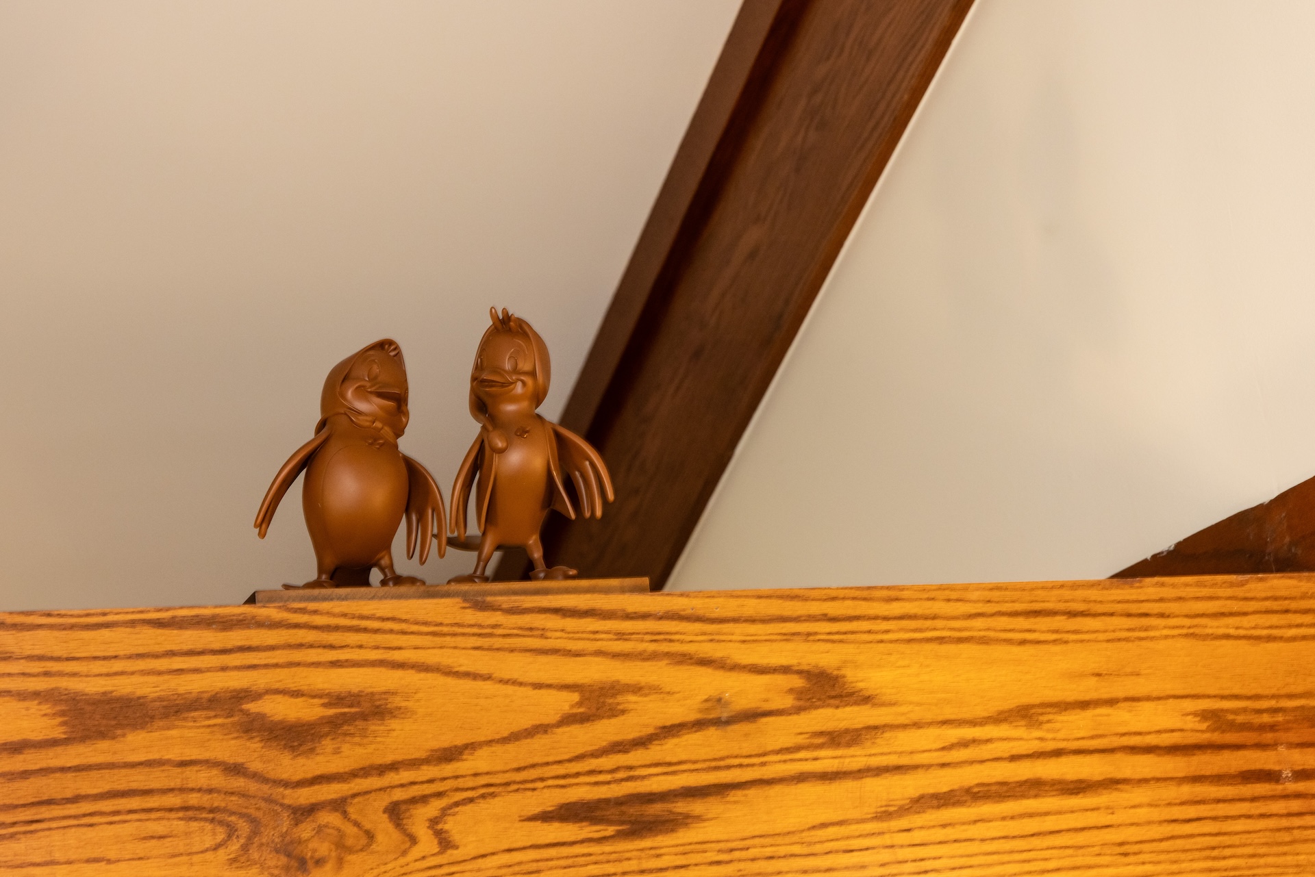 Two Brown Cartoon Duck Figurines Perched on a Wooden Shelf Against a Beige Wall with a Curved Dark Wood Beam in the Background