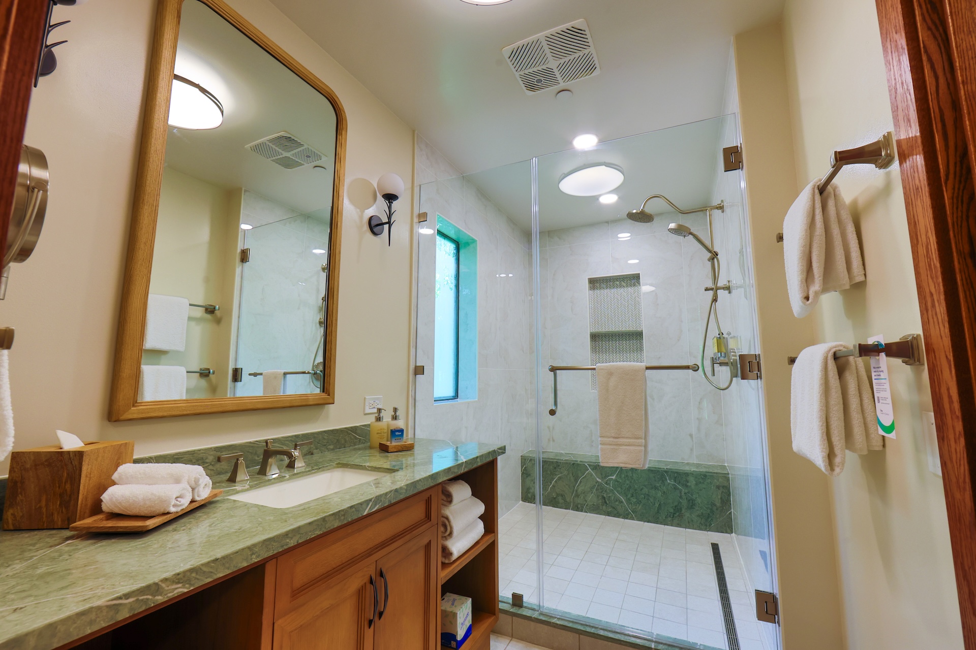 Bathroom with Green Marble Countertop Wooden Vanity and a Large Framed Mirror Glass Shower Enclosure with Built in Shelves and Multiple Towels on Racks