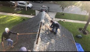 Two workers on a sloped roof repair the ridge with ropes and tools. The scene suggests teamwork and safety awareness, set in a bright residential area.
