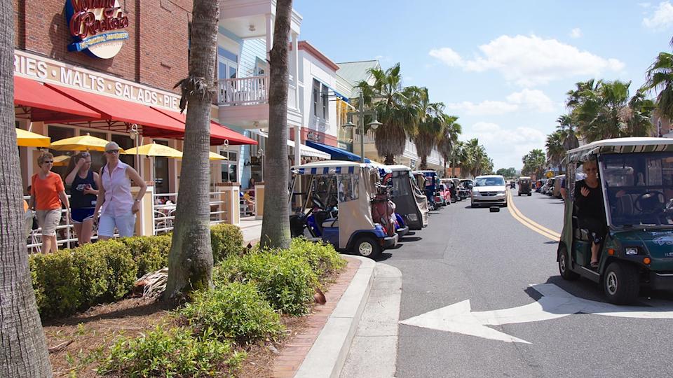 A wide outdoor shot of a busy street in a sunny town, lined with numerous golf carts parked on both sides. On the left, people walk on a sidewalk in front of commercial buildings with red awnings, including a 'Johnny Rockets' restaurant. Lush green bushes and palm trees are prominent in the foreground and along the street. In the foreground on the right, a dark green golf cart with a woman smiling and waving from the driver's seat is positioned as if turning onto the main road, marked with a large white arrow on the asphalt. Other cars are visible in the distance on the two-lane road under a bright blue sky with scattered clouds.