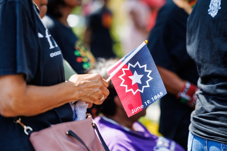 Someone holds the Juneteenth flag, featuring a blue upper half, a red lower half, and a downward-curved arc with a centered, white five-pointed star surrounded by a starburst. the bottom of the flag includes the text "June 19, 1865."