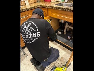 A skilled technician from Plumbing Around the Clock is pictured performing a precise under-sink repair in a residential kitchen. Wearing the company