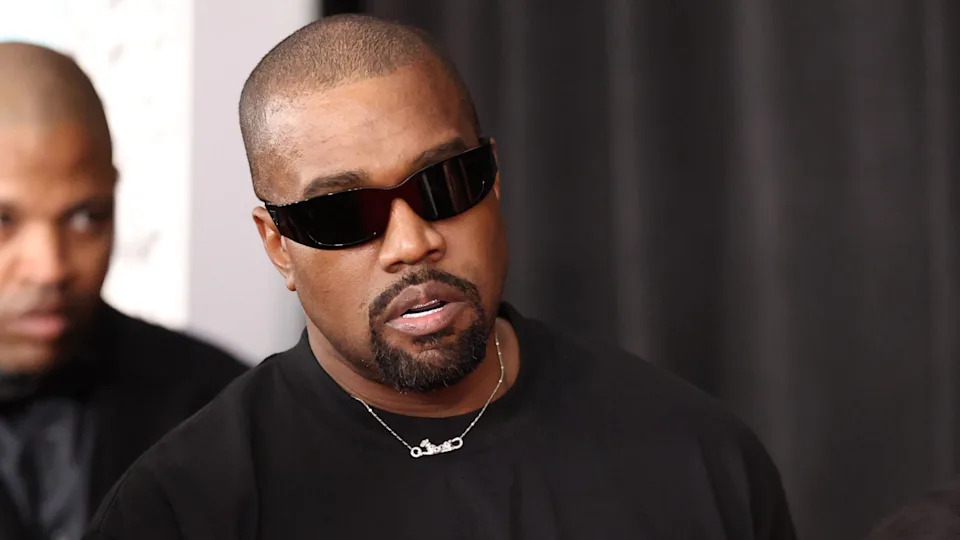 Kanye West wearing sunglasses, a black shirt, and a necklace, at an event with a black backdrop.