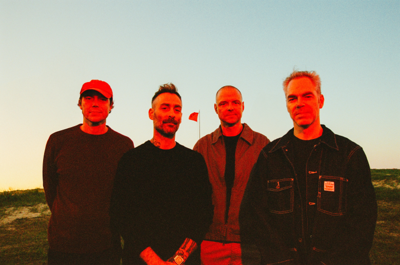 A medium-shot, outdoor band portrait of American Football during a vibrant sunset. From left to right, the members are Steve Holmes, Mike Kinsella, Steve Lamos, and Nate Kinsella. They are standing in a horizontal line, facing the camera with neutral expressions. They are dressed in casual, dark-toned clothing against the backdrop of an open landscape under a clear, pale-blue to orange-gradient sky. A strong, warm-toned red light source is directed from the side, casting the band members in dramatic, saturated red and orange hues while creating deep shadows.