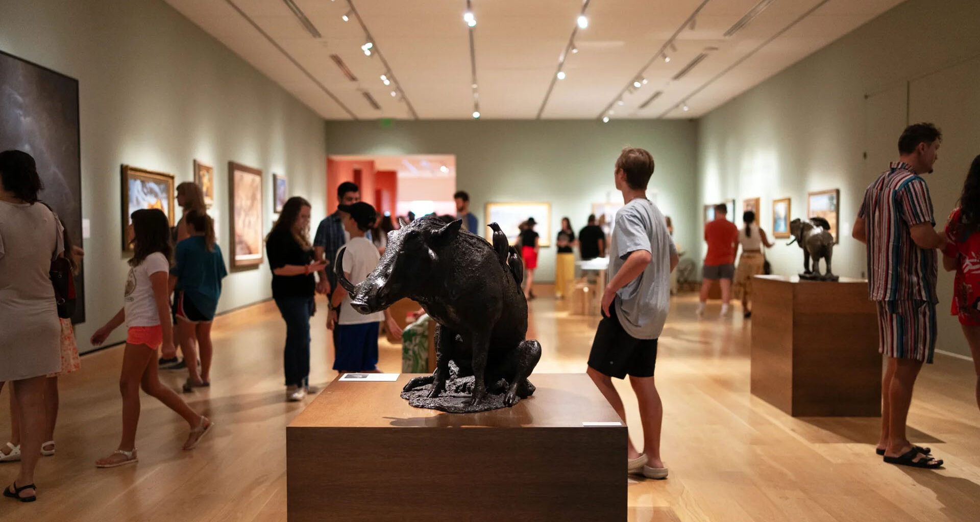 An art gallery interior featuring visitors observing sculptures and paintings displayed along the walls and on pedestals.