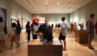 An art gallery interior featuring visitors observing sculptures and paintings displayed along the walls and on pedestals.
