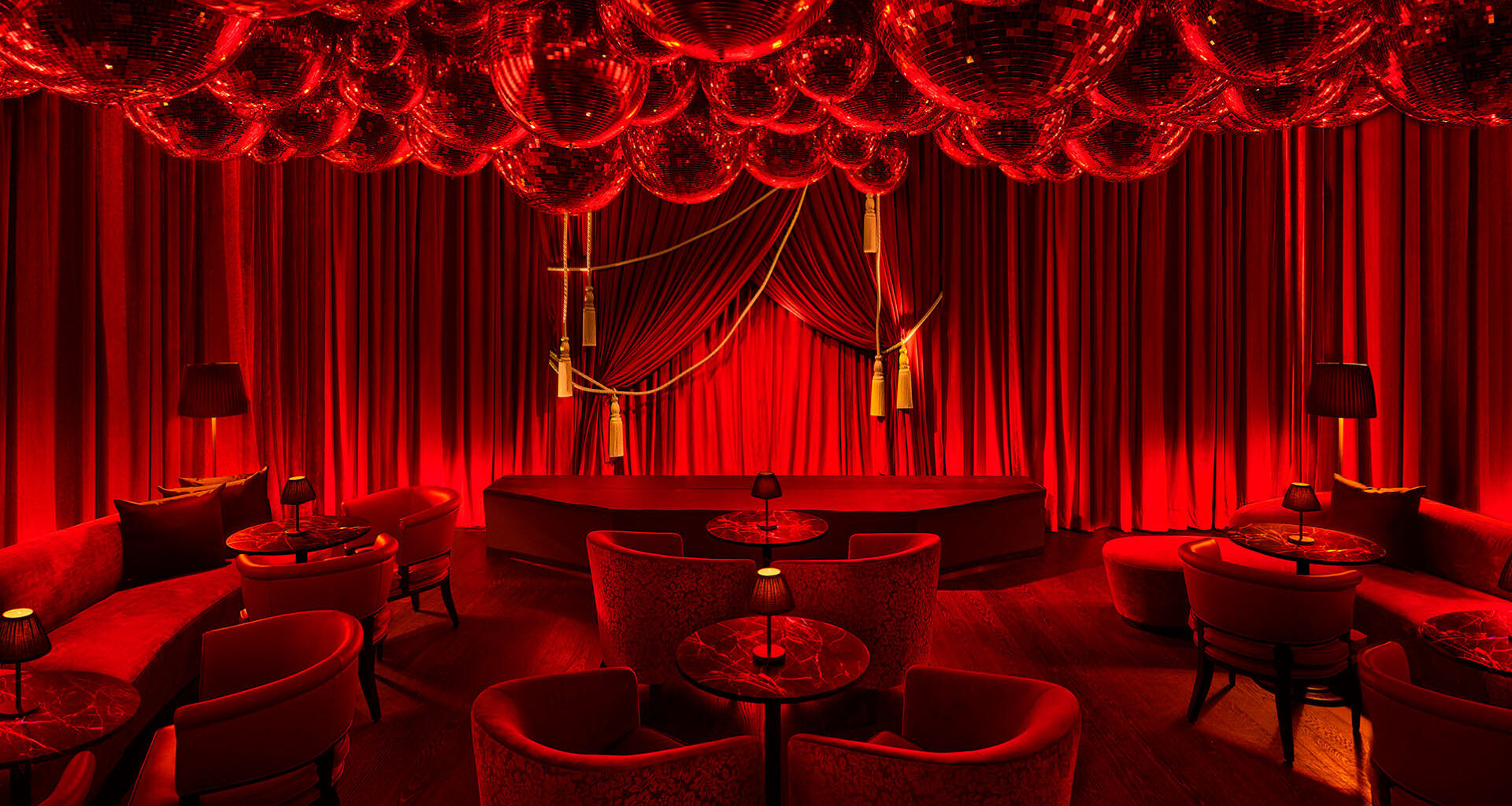 A luxurious lounge interior featuring deep red lighting, velvet curtains, plush seating arranged around small round tables, and a ceiling filled with disco balls.