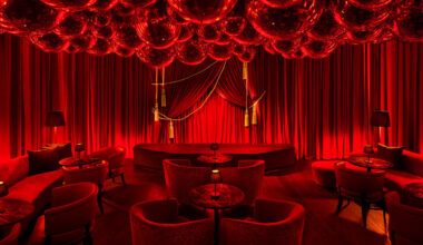 A luxurious lounge interior featuring deep red lighting, velvet curtains, plush seating arranged around small round tables, and a ceiling filled with disco balls.