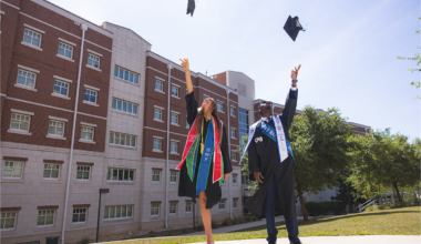 Two graduates throw their graduation caps in the air in front of a dormitory.