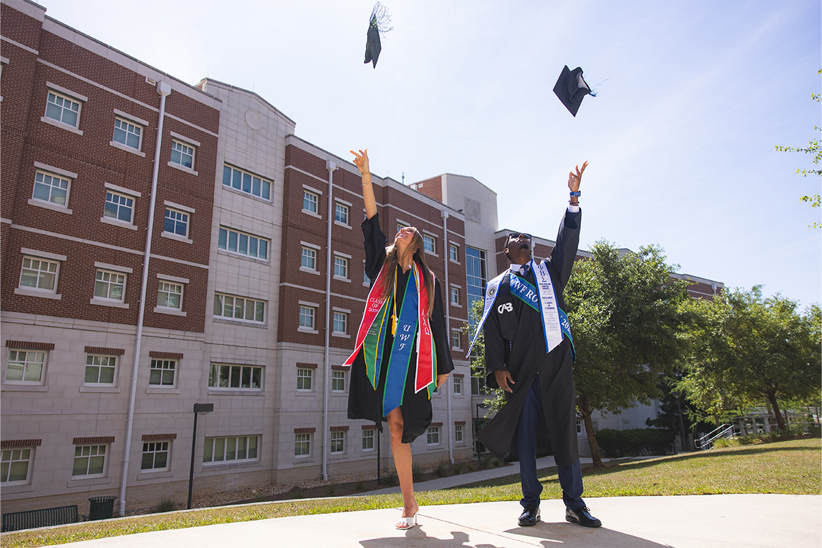 Two graduates throw their graduation caps in the air in front of a dormitory.