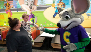 Toddler held by an adult reaching out to touch Chuck E. Cheese in a family entertainment center.