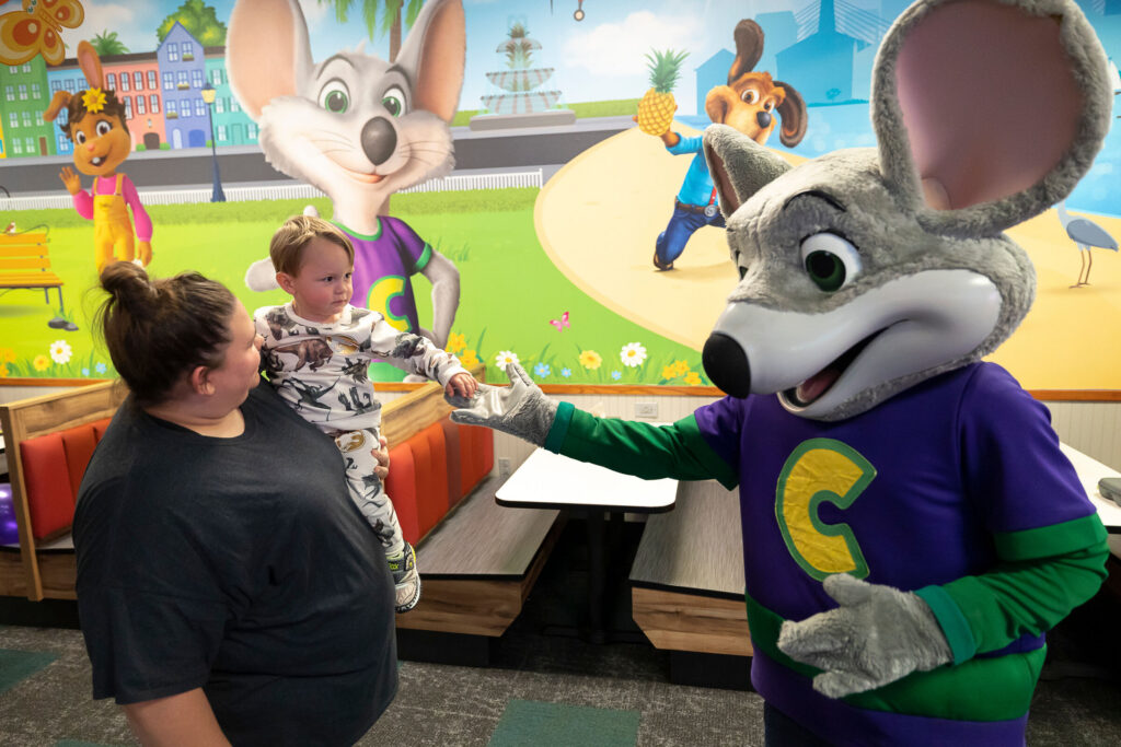 Toddler held by an adult reaching out to touch Chuck E. Cheese in a family entertainment center.