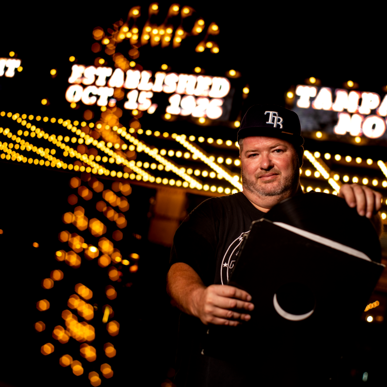A portrait of DJ Sandman standing at night outside the historic Tampa Theatre in Tampa, Florida. The individual is wearing a black shirt and a Tampa Bay Rays baseball cap while holding a vinyl record sleeve. In the background, the glowing marquee lights display "Established Oct. 15, 1926.