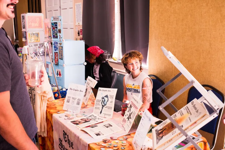A smiling person at a table displaying copies of the Sapphic Sun at New Moon Makers' Market
