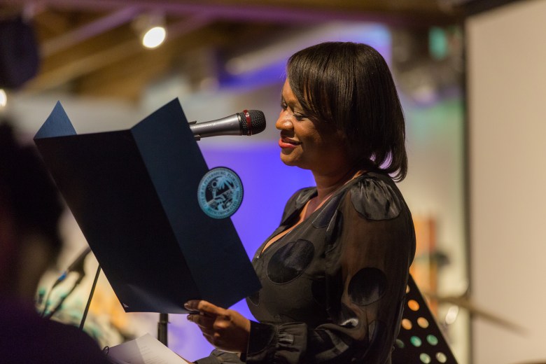 A medium-shot, indoor candid photograph of Dr. Kanika Tomalin speaking at an event inside the Studio@620 in St. Petersburg, Florida. Tomalin is positioned to the right, looking toward the left with a warm, engaged smile. They are wearing a long-sleeved, dark-colored, semi-sheer black top with a subtle circular pattern. They are holding a dark, open folder or script in their hands. A professional studio microphone on a stand is positioned in front of them. The background is softly out of focus, showing the interior of the venue with hints of studio lighting and a neutral, professional environment.