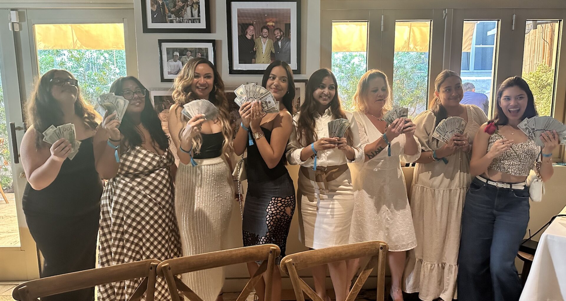 photo of eight women posing together in a row holding up dozens of $100-dollar bills