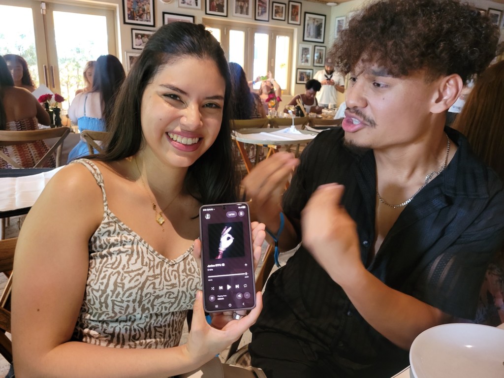 Photo of a young woman and man posing at a table. The woman holds up her phone, with the screen showing Drake's "Janice STFU" streaming on Apple Music