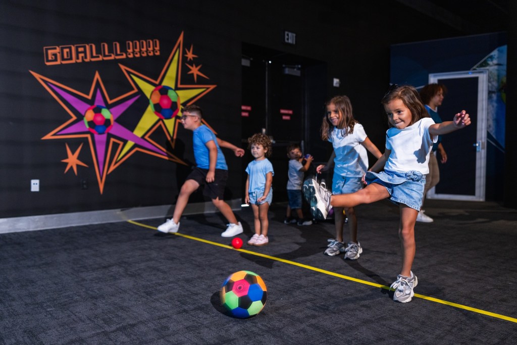 photo of children inside a museum kicking a soccer ball behind a yellow line of tape on the floor