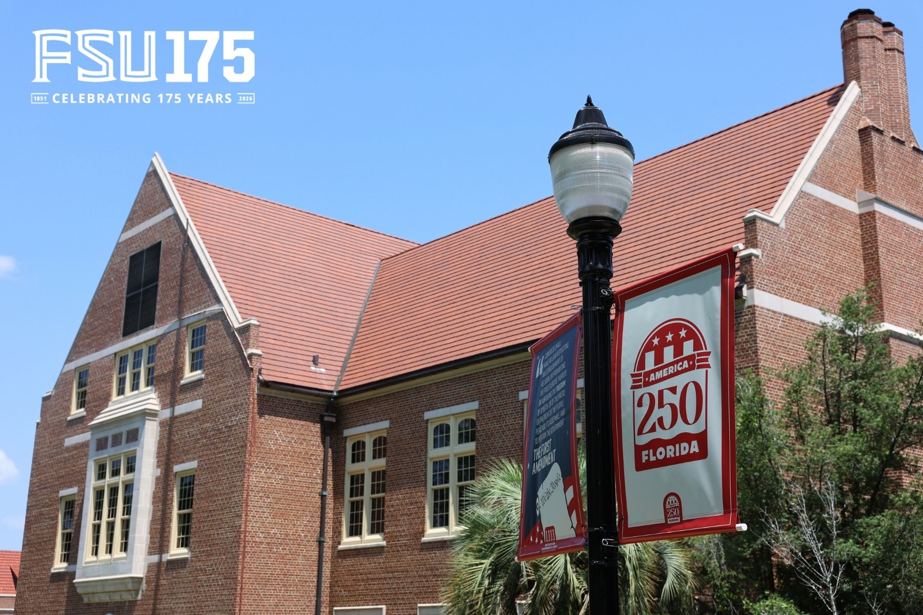 A brick building at Florida State University stands under a clear blue sky, partially framed by a lamp post featuring commemorative red and white banners.