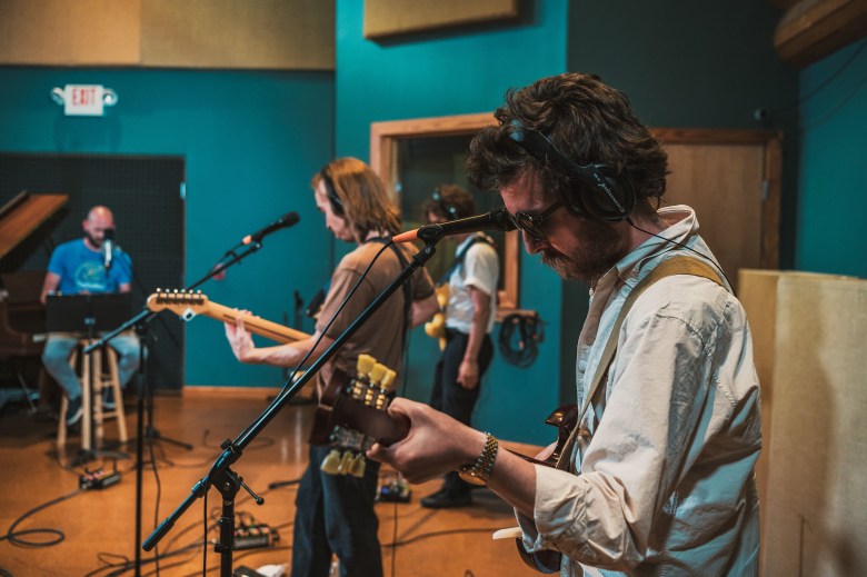 A wide, eye-level, live performance photograph of the band Fast Preacher recording at WMNF in Tampa, Florida. In the foreground, a guitarist with wavy brown hair, wearing a cream-colored button-down shirt and headphones, plays an electric guitar while facing toward the center of the room. To their left, another band member wearing a tan t-shirt plays a separate electric guitar. In the background, a third band member is seated at a piano, also wearing headphones and playing into a studio microphone. The studio features teal-colored walls, wood flooring, and an 'EXIT' sign illuminated in red above a doorway in the background.
