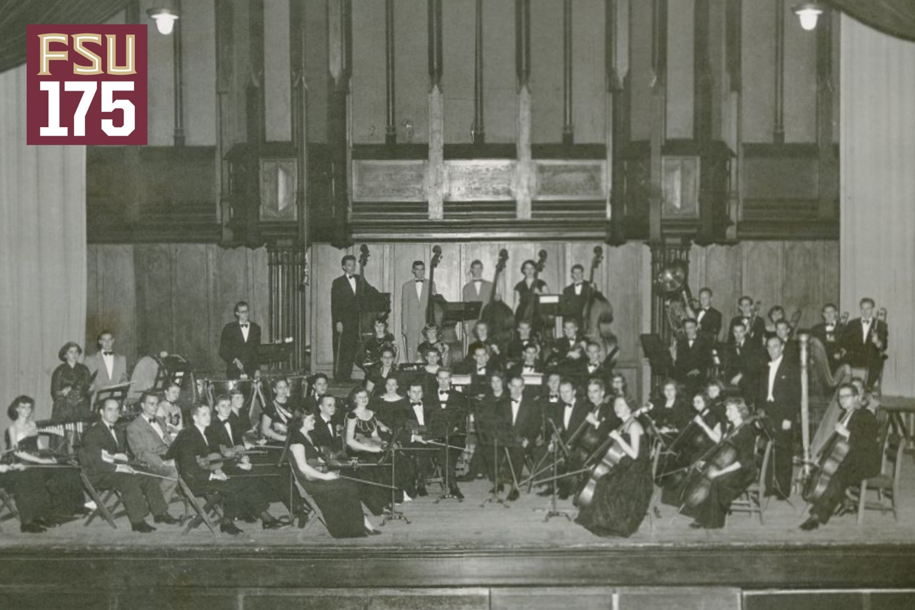 An archival, black-and-white photograph shows a large student orchestra posing with their instruments on a wooden stage, set against a background of prominent organ pipes and a garnet-and-gold "FSU 175" logo in the upper left corner.