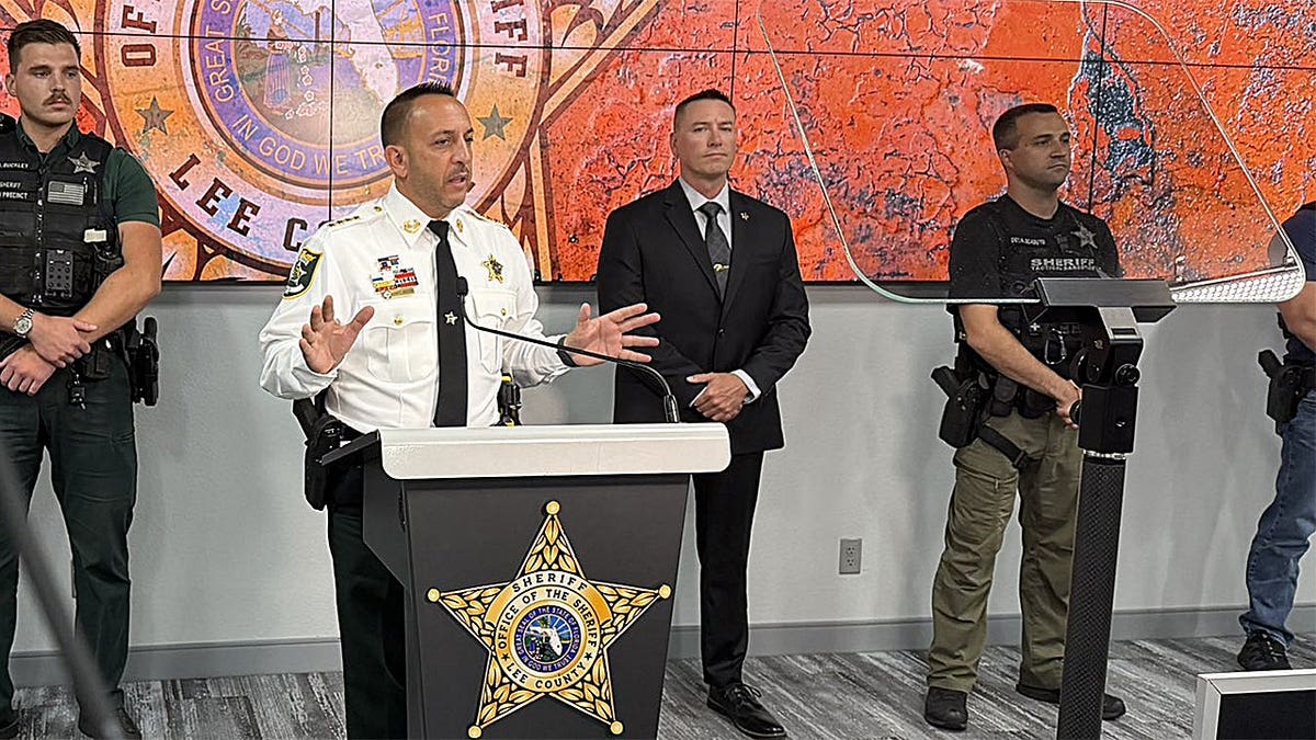 Lee County Sheriff Carmine Marceno speaks at a podium during a press conference while deputies and investigators stand behind him.