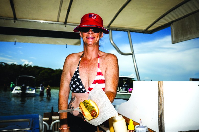 Woman wearing an American flag bikini top smiles and holds out a hot dog onboard Nauti Dogs hot dog boat.