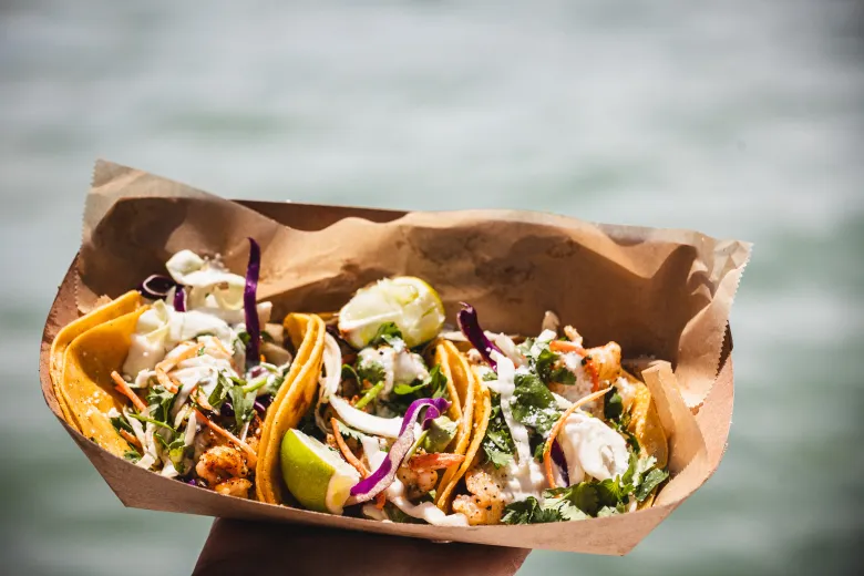 A colorful paper basket of tacos from Tiki's Tacos held up against a watery background.