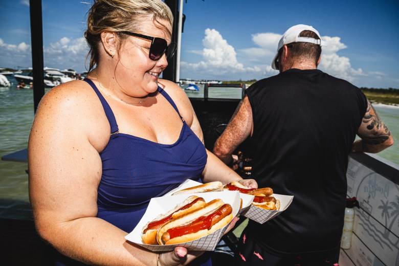 A woman holding several hot dogs in front of a man grilling on a food boat, Beachin' Bites