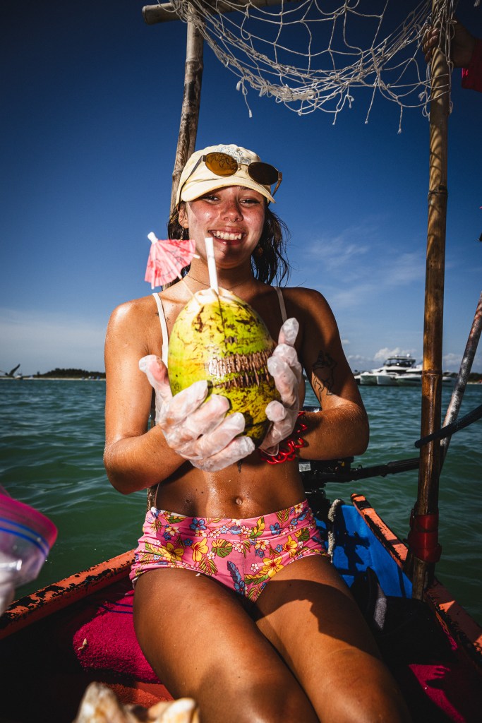 Smiling woman in a bikini wearing rubber gloves holds up a coconut with a straw in it aboard the Fruit Float