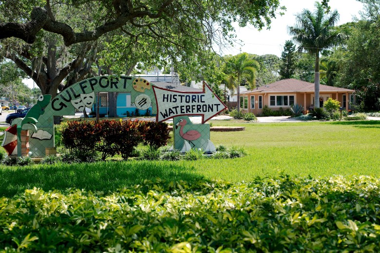 A sunny outdoor photograph of the colorful "Historic Waterfront" welcome sign in Gulfport, Florida. The artistic sign, featuring a painted arrow, a pink flamingo illustration, and theatrical masks, sits on a lush green lawn surrounded by mature trees, landscaping, and tropical palm trees. A residential building is visible in the background under a bright blue sky.