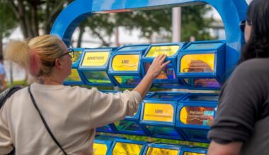 A woman in a light sweater interacts with a blue, interactive, word-display installation outdoors. Her expression is thoughtful and engaged.