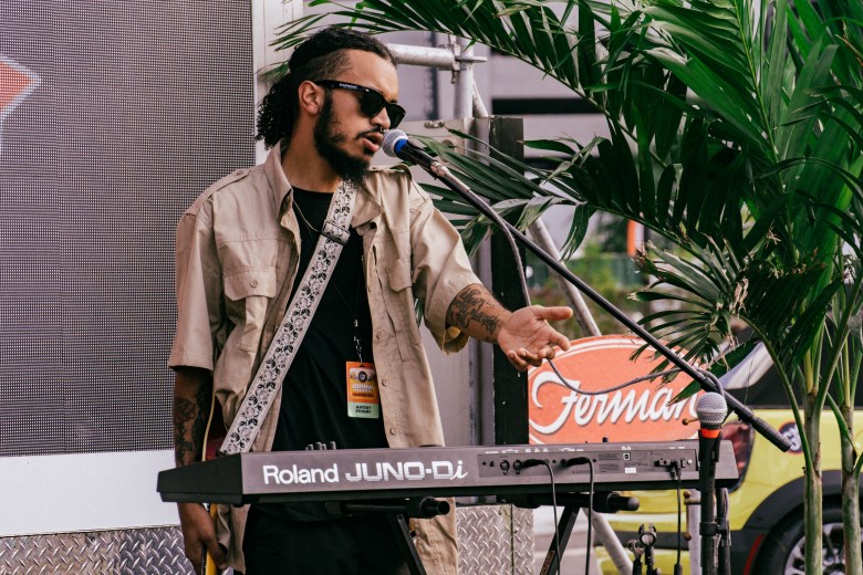 Musician Javier Solaris performs live on stage during the Gasparilla Music Festival at Meridian Fields in Tampa, Florida. The artist, wearing sunglasses, a beige short-sleeved button-up shirt over a black t-shirt, and a festival credential lanyard, stands behind a Roland JUNO-Di electronic keyboard. They are singing into a microphone with one hand extended outward, surrounded by tropical palm fronds and stage equipment under daylight.