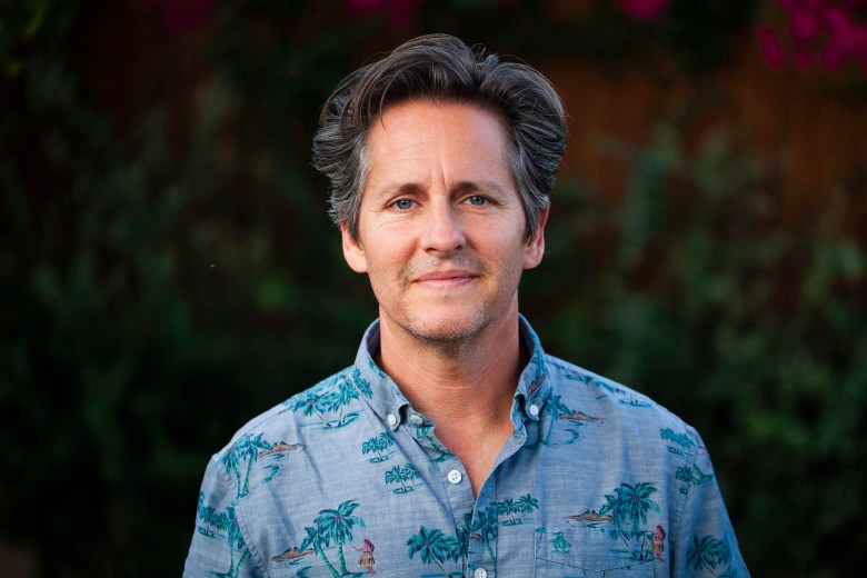 A medium-shot outdoor portrait photograph of singer-songwriter Josh Rouse. They have short, swept-back graying dark hair and are wearing a light blue, short-sleeved button-down shirt featuring a tropical palm tree pattern. They are looking directly at the camera with a soft, relaxed smile. The background is softly out of focus, showing dark green foliage and hints of bright pink flowers under soft, natural lighting.