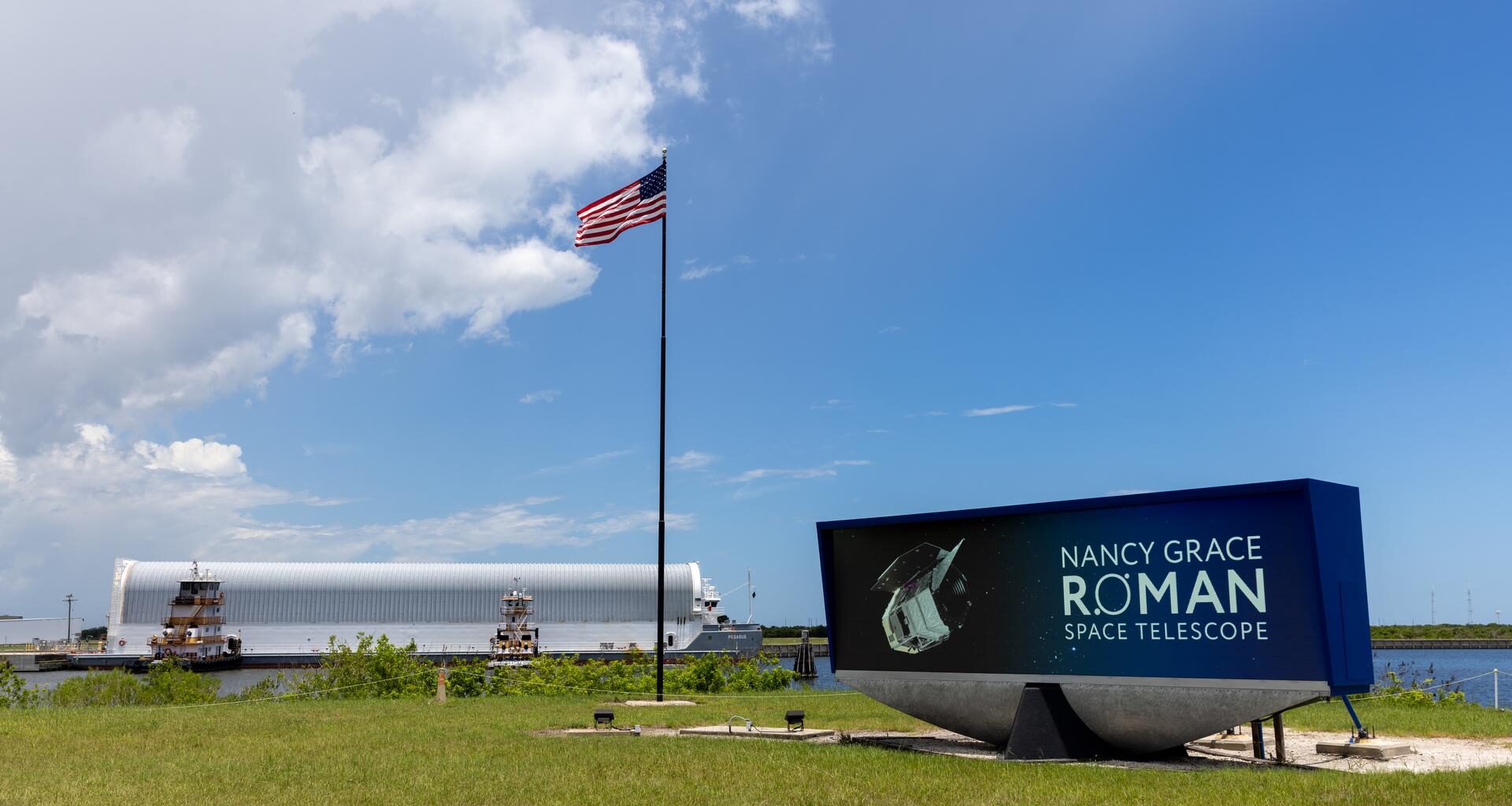 Image shows Nancy Grace Roman Telescope insignia on the countdown clock near an American flag on Sunday, June 21, 2026 at NASA's Kennedy Space Center in Florida. In the background is a white cylinder on a barge carrying NASA's Nancy Grace Roman Space Telescope with blue skies and a few puffy clouds.