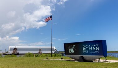 Image shows Nancy Grace Roman Telescope insignia on the countdown clock near an American flag on Sunday, June 21, 2026 at NASA's Kennedy Space Center in Florida. In the background is a white cylinder on a barge carrying NASA's Nancy Grace Roman Space Telescope with blue skies and a few puffy clouds.