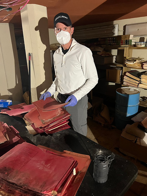 A man wearing a white face mask, black baseball cap, and blue protective gloves stands behind a table stacked with old red legal files in a dimly lit basement or storage room.