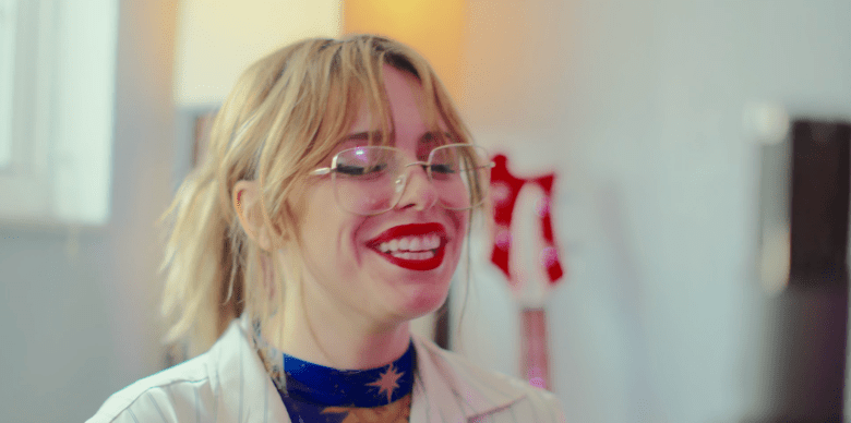A close-up, candid video-still portrait of chef Nicole Hoffer. They are smiling broadly and looking slightly off-camera with a joyful expression. They have blonde hair pulled back into a ponytail with loose bangs framing their face. They are wearing large, transparent-frame glasses, bright red lipstick, and a white chef's coat. A dark-blue scarf with a gold star pattern is visible around their neck. The background is softly blurred, showing a bright, neutral-toned interior kitchen environment.