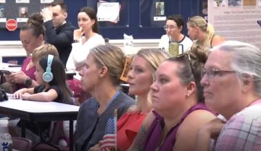 A crowd of parents sits at desks, questioning the School Board about a coach who was arrested.