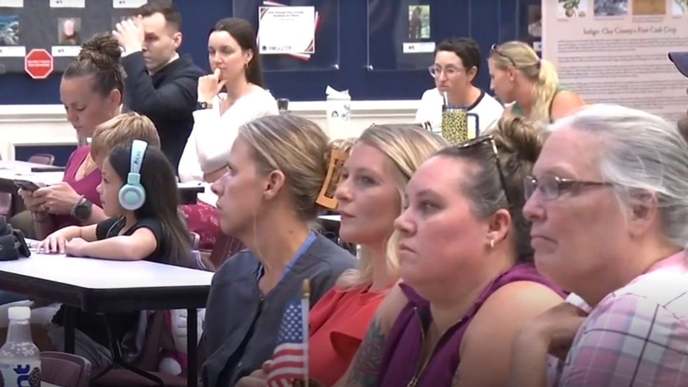 A crowd of parents sits at desks, questioning the School Board about a coach who was arrested.