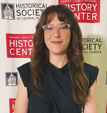 Photo of Rachel Williams, historian. Woman with long medium brown hair, clear glasses, and black top standing in from of step and repeat banner.
