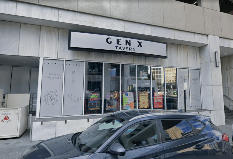 An eye-level exterior photograph of the Gen X Tavern storefront in downtown Tampa. The facade features a modern, neutral-toned building with a prominent, rectangular sign reading 'GEN X TAVERN' in clean, dark, sans-serif lettering. Below the sign is a large, glass-paneled entrance with several posters and menu displays visible inside the windows. The building's exterior is characterized by light gray architectural paneling and clean, industrial lines.