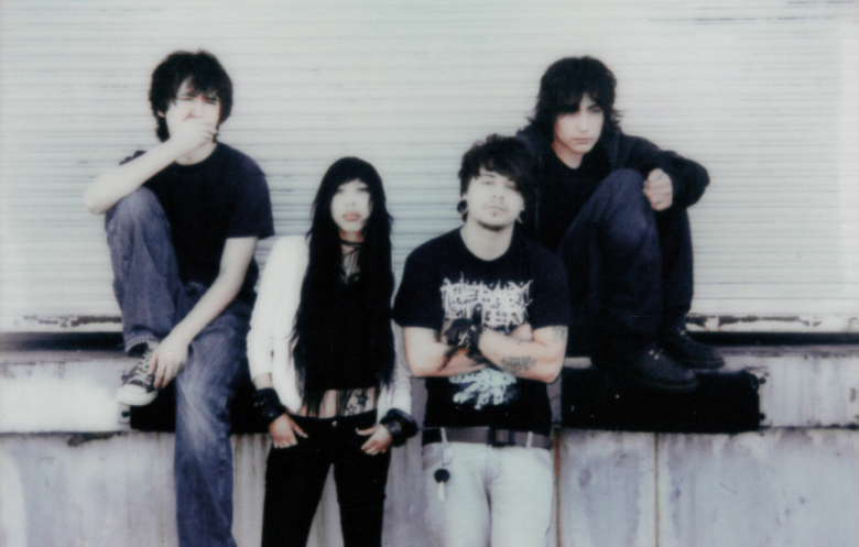 A lo-fi, vintage-style promotional photograph of the skramz band Septemberistheonlytimeidontthinkofyou. Four band members are posed casually against a white, corrugated rolling garage door or shipping container. Two individuals sit elevated on a ledge, while the other two stand directly in front. The group is dressed in a dark, alternative aesthetic featuring plain t-shirts, a graphic metalcore band shirt, black pants, and a white outerwear layer.