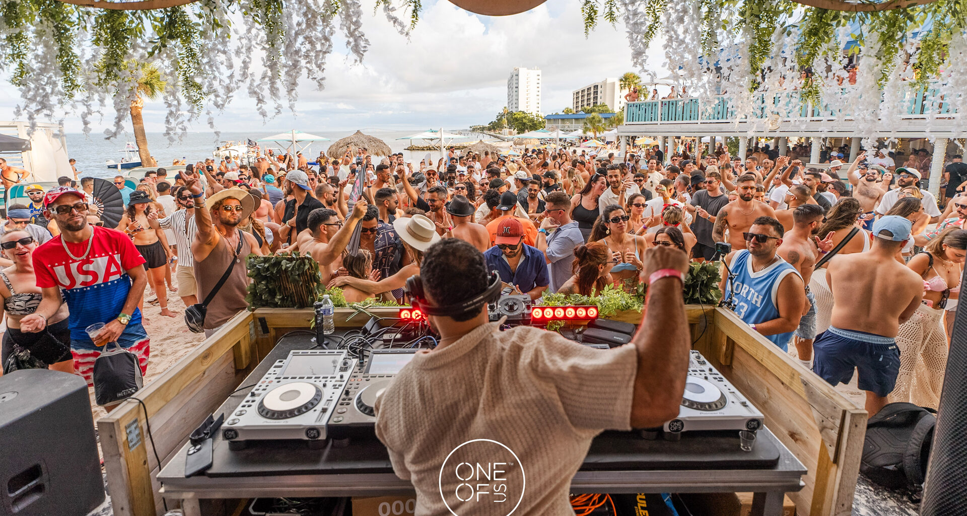 A beach party featuring a DJ performing on a wooden deck with turntables and a mixer, facing a large crowd of people dancing in front of palm trees and beach umbrellas.
