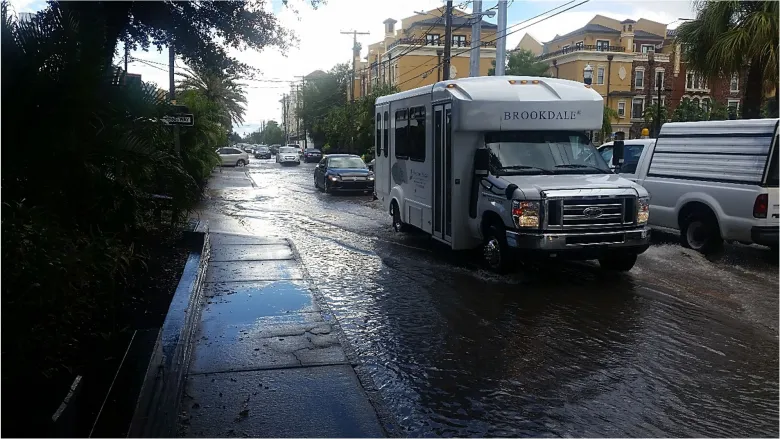 A flooded street view of South Howard Avenue near West Bristol Avenue in Tampa, Florida, during September 2016. High, murky floodwaters cover the entire roadway and parts of the concrete sidewalk, reflecting the sky. In the center, a white Brookdale shuttle bus and a white pickup truck wade slowly through the deep water, while other vehicles are visible lined up further down the flooded street under an overcast sky.