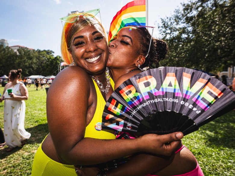 A smiling sapphic couple embraces at St. Pete Pride. One of them holds a hand fan with "be brave" written in rainbow letters.