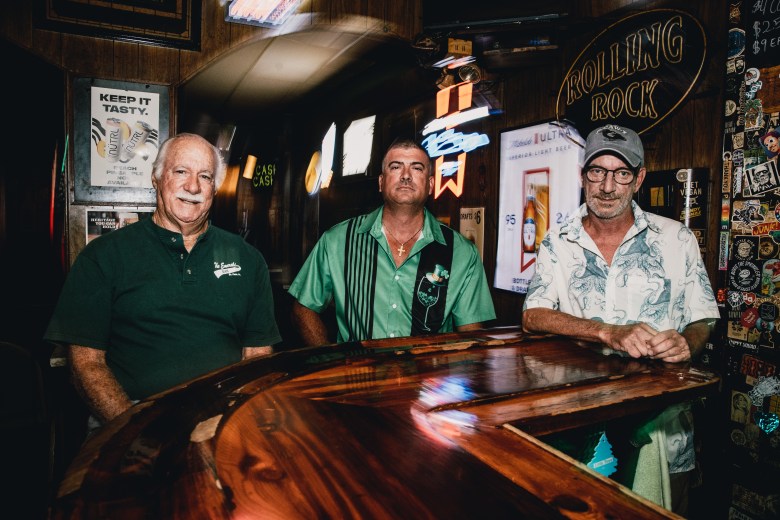 A medium-shot, indoor photograph taken inside the Emerald Bar in St. Petersburg, Florida. Three people sit at the bar's signature polished, curved wooden counter. From left to right: George Marano, Michael Marano, and Mikey Brinkmann. George Marano is wearing a dark green polo shirt; Michael Marano is in the center wearing a light green button-down shirt; and Mikey Brinkmann is on the right wearing a short-sleeved, patterned white shirt and a gray baseball cap. The background features the dimly lit, wood-paneled interior of the bar, adorned with vintage 'Rolling Rock' signage and various memorabilia on the walls.